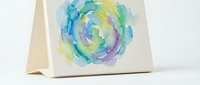 Watercolor spiral swirl in vibrant blue, green, yellow and purple tones on folded cream cardstock. Suitable for greeting cards, stationery and artistic designs.