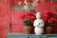 White Buddha statue with red flowering plants in terracotta pots on rustic wooden shelf against weathered red wall for meditation and zen decor.