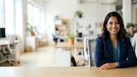 Young South Asian woman in professional navy blazer and teal top smiling at desk in modern office environment with blurred colleagues and plants, suitable for corporate, business, and leadership concepts.
