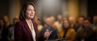 Professional businesswoman speaking at podium during corporate conference with audience in background for leadership presentations.