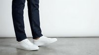 Legs of a person wearing dark blue jeans and clean white sneakers standing on a concrete floor against a light gray wall.