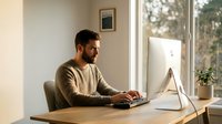 Young bearded man working on computer at wooden desk by window in bright modern home office setup for remote work and productivity.