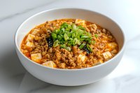 Spicy mapo tofu with minced meat and green onions in white ceramic bowl on marble surface, traditional Chinese Sichuan dish garnished with fresh herbs.