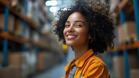 Young African American woman with natural curly hair and bright smile wearing yellow shirt standing in warehouse storage facility with shelves background.