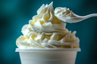 Creamy swirled soft serve vanilla ice cream in white cup with spoon against turquoise background, macro food photography showing texture and peaks.