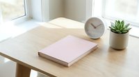 Blank pink notebook on wooden desk with clock and succulent plant by bright window, workspace organization and productivity concept.