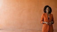Young African American woman in professional orange blazer standing against warm terracotta wall, suitable for corporate, business, and leadership marketing materials.