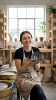 Young woman, Caucasian ethnicity, potter and ceramicist, smiling while holding handmade ceramic bowl in bright studio workshop with large windows, wooden shelves, plants, and pottery supplies.
