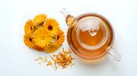 Calendula flower tea in glass cup with fresh yellow blooms and dried petals on white background, herbal remedy for wellness and natural health.