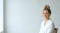 Blonde woman with hair in top bun, wearing white button-up shirt, sitting on wooden chair against white wall background, smiling at camera.
