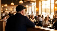 Senior man in black cap observing busy restaurant dining room with warm lighting and crowded tables during peak hours.