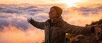 Mature man with brown skin wearing winter jacket and beanie standing on mountain peak above clouds at golden hour sunrise, arms outstretched in celebration and freedom.