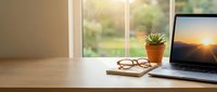 Modern home office workspace with laptop displaying sunset, eyeglasses and succulent plant on wooden desk near bright window for remote work productivity.