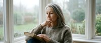 Mature woman with long gray hair and light skin tone sitting by large windows, wearing a cozy knit sweater, reading a book while gazing thoughtfully outdoors on an overcast day.