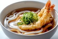 Tempura shrimp udon noodle soup in white ceramic bowl with chopped green onions and clear savory broth, traditional Japanese cuisine close-up photography.