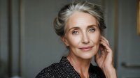 Elegant mature woman with silver hair and natural makeup touching face gently against neutral background, portraying confidence and grace.