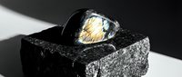Polished black obsidian sphere with golden rutile inclusions resting on textured volcanic rock surface with dramatic lighting and shadows.
