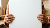 Mature adult with medium skin tone holding blank white paper sheet for mockup, advertising, or promotional messaging in indoor setting.