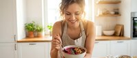 Young Caucasian woman enjoying healthy breakfast bowl with berries and granola in bright modern kitchen. Nutritious morning meal lifestyle concept.