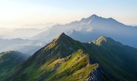 Majestic mountain ridge illuminated by morning sunlight, showcasing dramatic peaks and valleys with lush green slopes against misty distant mountains in summer landscape.