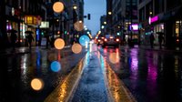 Rainy city street at night with colorful bokeh lights reflecting on wet asphalt. Urban evening scene with blurred traffic and illuminated storefronts.