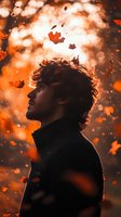 Young man silhouette in profile against autumn sunset sky with falling maple leaves and bokeh lights creating magical atmospheric mood.