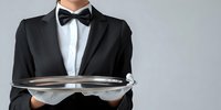 Professional waiter in formal black tuxedo and bow tie holding empty silver serving tray against gray background, ready for luxury restaurant service.