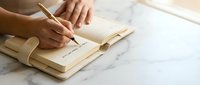 Woman writing in wooden notebook with golden pen on marble surface for journaling and creative planning goals.