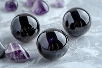 Polished black obsidian spheres with purple amethyst crystals on white marble surface for healing meditation and spiritual wellness practices.