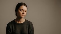 Young Caucasian woman with closed eyes in contemplative pose wearing dark sweater against neutral beige background for mental health awareness campaigns.