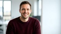 Confident middle-aged Caucasian businessman smiling in modern office wearing burgundy sweater for corporate headshot and professional marketing materials.