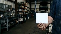 Mechanic in work uniform holding blank white card in industrial garage workshop with tools and equipment on shelves in background.