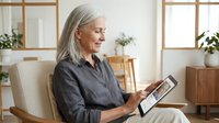 Senior woman with gray hair using digital tablet while sitting in modern living room chair, smiling as she reads or browses online content at home.