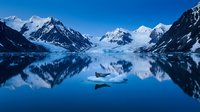 Serene alpine lake surrounded by snow-capped mountains and evergreen forests, with small boats floating on calm blue water reflecting the winter landscape.