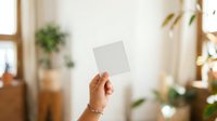 Female hand holding blank white card in bright interior with plants and natural light, suitable for mockups, branding, and promotional materials.