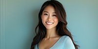 Young Asian woman with long dark wavy hair and bright genuine smile wearing light blue top against turquoise wall background, expressing joy and confidence in casual portrait.