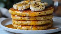 Stack of fluffy poppy seed pancakes topped with fresh banana slices and drizzled with golden maple syrup on white ceramic plate, close-up food photography.