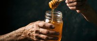 Elderly hands holding glass jar while drizzling golden honey with wooden dipper against dark background for natural food and wellness concepts.