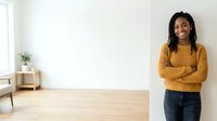 Young Black woman with braided hair wearing mustard yellow sweater and dark jeans, standing confidently with crossed arms in bright minimalist interior space with wooden flooring.