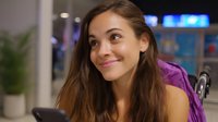 Young Mediterranean woman with long brown hair smiling warmly while leaning forward, wearing purple top, expressing genuine happiness and positive emotions in modern interior setting.