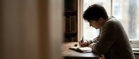 Young man writing in notebook by window in cozy room with warm natural light creating peaceful study atmosphere for journaling or creative work.