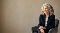 Mature woman with long gray curly hair wearing dark blazer and white shirt, seated in chair against neutral beige background, professional portrait for business and corporate use.