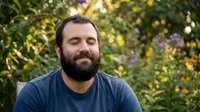 Portrait of a bearded man with dark hair and facial hair wearing a navy blue shirt, posed outdoors against a blurred garden background with yellow and purple flowers.