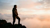 Young man in dark jacket standing on mountain peak above clouds at sunrise, contemplating horizon. Represents achievement, adventure, and personal growth for motivational and lifestyle content.