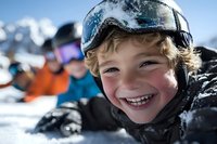 Cheerful young Caucasian boy wearing ski goggles and winter gear smiles brightly during snow sports activity with blurred skiers in background on sunny mountain slope.