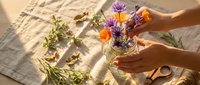 Woman arranging fresh purple and orange flowers in glass vase with herbs on linen cloth, natural sunlight, floral design and craft project.