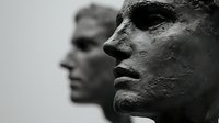 Dramatic profile view of two sculptural faces in dark monochrome showing textured surface detail and emotional expression against blurred background.