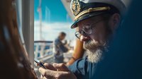 Experienced sea captain with white beard using smartphone on ship bridge while navigating maritime vessel during sunny day at sea.