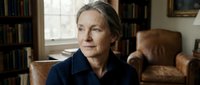 Mature Caucasian woman in navy shirt sitting thoughtfully in cozy home library with leather armchair and bookshelves, natural window light creating warm atmosphere.