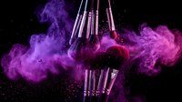 Professional makeup brushes with vibrant purple powder explosion on black background. Cosmetic tools surrounded by colorful dust cloud for beauty industry.
